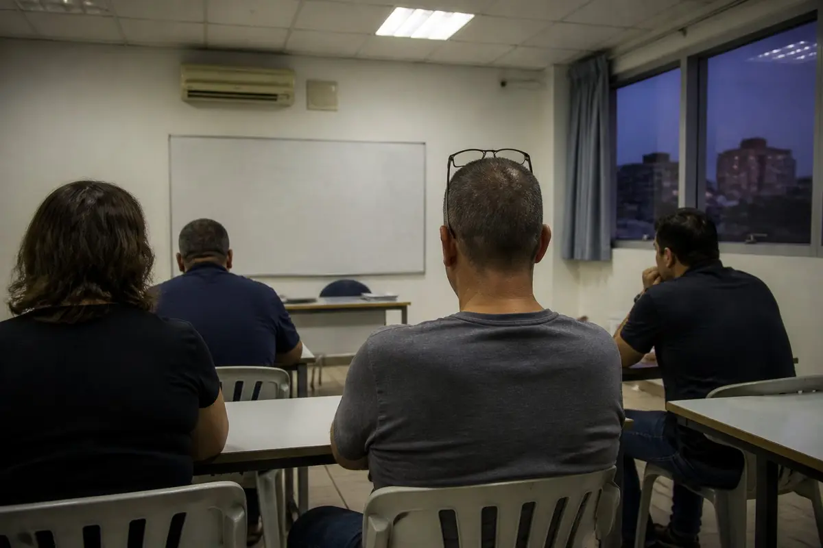 כיתת ערב במכללה, כמה מבוגרים יושבים בגבם למצלמה מול לוח לבן ריק, ובחלון בניינים בדמדומים