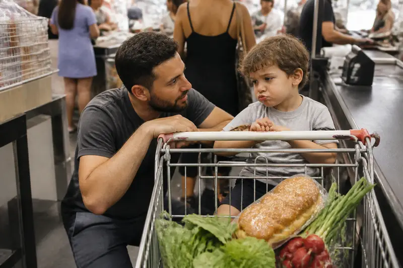 אב כורע ליד עגלת קניות בתור לקופה בסופר ומדבר בשקט עם בנו הקטן שיושב בעגלה ברוגז
