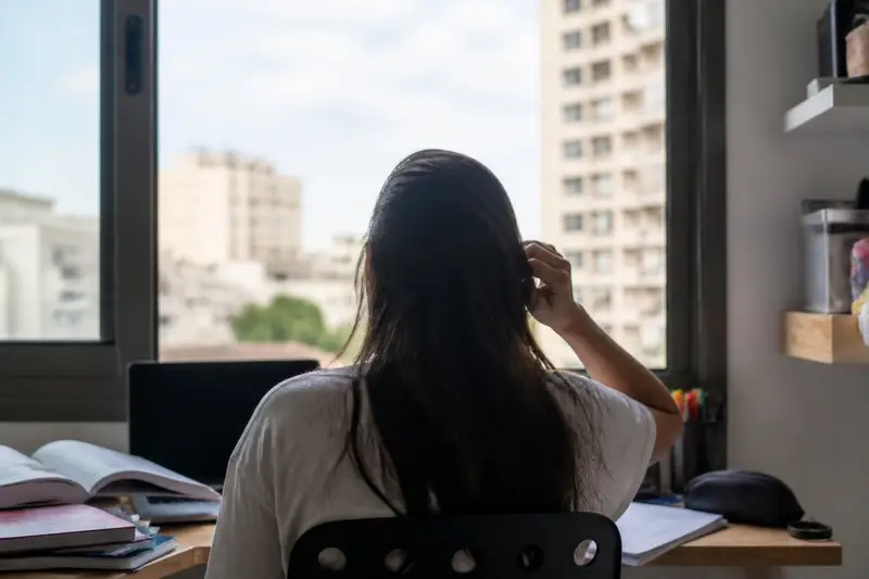 נערה יושבת ליד שולחן לימוד מול חלון, מצולמת מאחור, יד אחת מורמת אל השיער