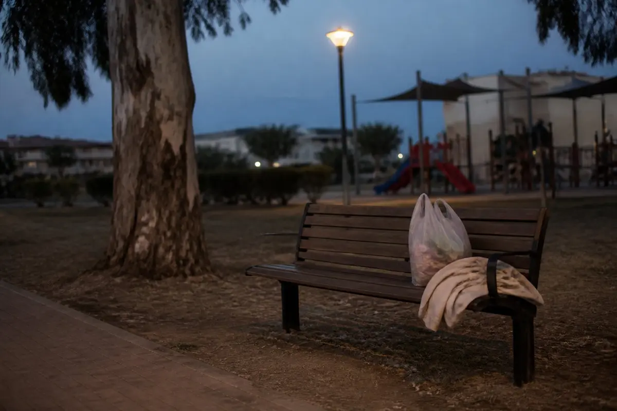 ספסל עץ בגינה שכונתית בשעת דמדומים, עם שקית קניות ומעיל מקופל בקצה אחד שלו