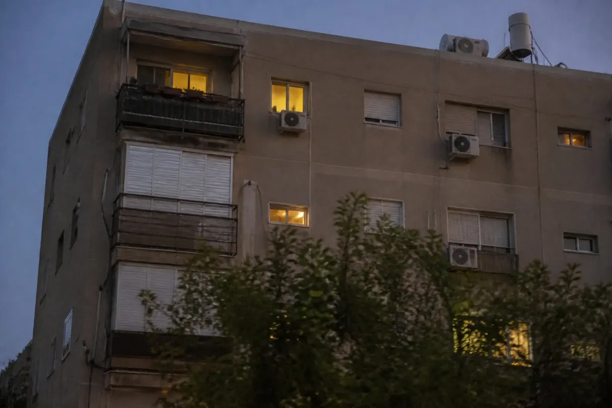 בניין מגורים ישראלי בשעת ערב, כמה חלונות מוארים באור צהוב ואחרים חשוכים, תריסים חצי סגורים ודודי שמש על הגג