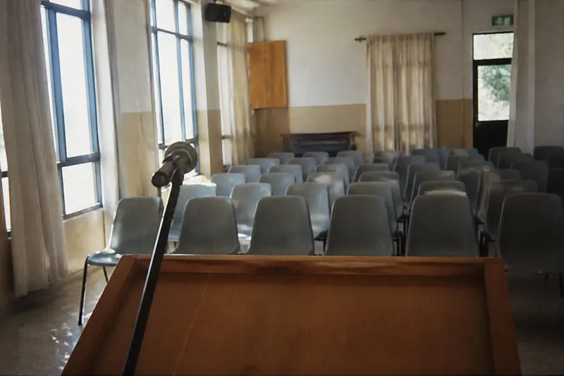 דוכן נואם מעץ עם מיקרופון, ומולו שורות של כיסאות פלסטיק אפורים ריקים באולם קהילתי באור בוקר