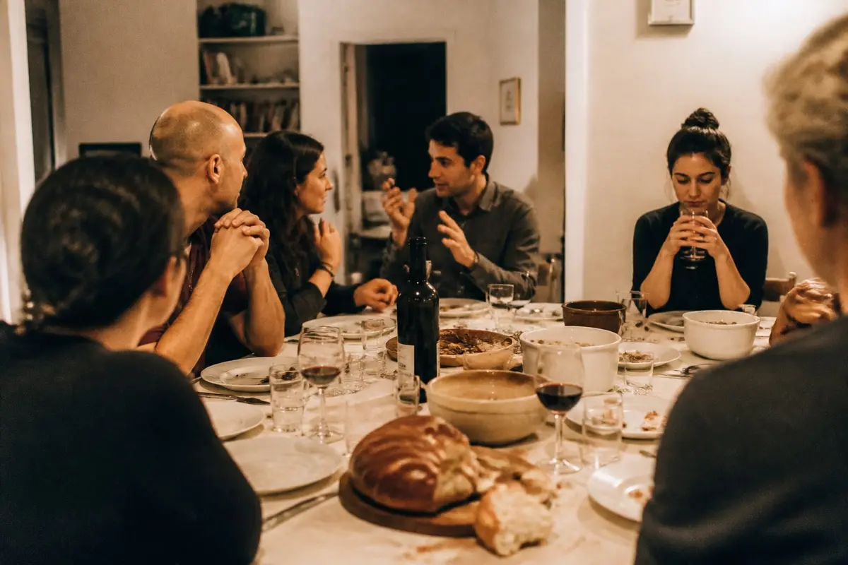 שולחן ארוחת שישי משפחתית, כמה מבוגרים משוחחים ואישה אחת יושבת מעט בצד, שקטה, ידיה סביב כוס