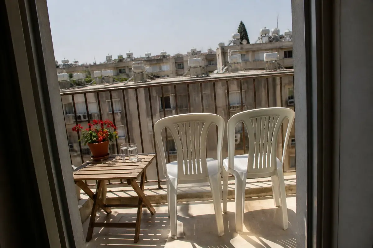 שני כיסאות באותו כיוון, ורבע שעה בלי טלפון. גם זו שפה. מרפסת קטנה בצהריים: שני כיסאות פלסטיק לבנים זה לצד זה מול נוף של גגות עם דודי שמש, ושתי כוסות מים על שולחן מתקפל