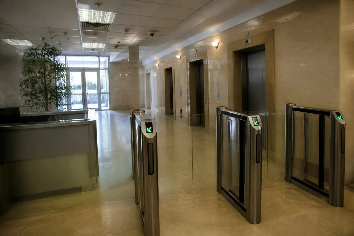 לובי של בניין משרדים בצהריים: קרוסלות כניסה עם קוראי כרטיסים, שורת מעליות ועמדת שומר ריקה