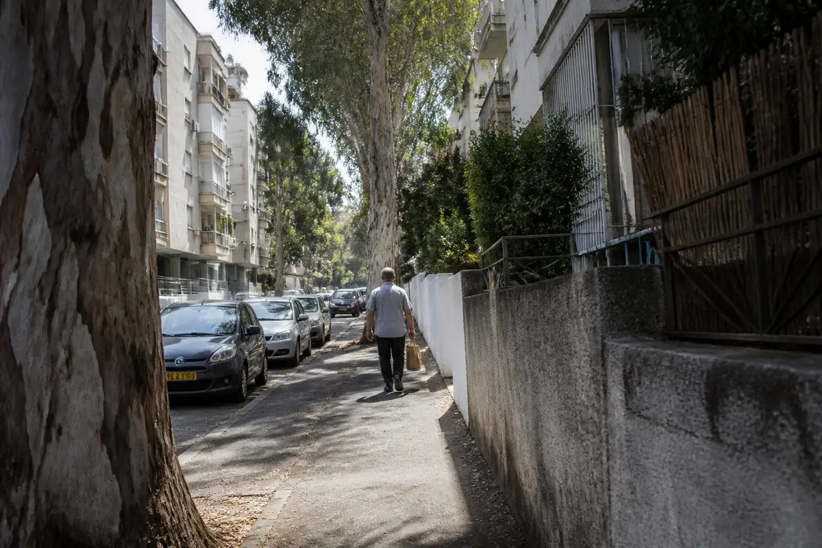 רחוב מגורים בצהריים: גבר הולך הרחק מהמצלמה עם שקית אוכל קטנה, בין מכוניות חונות ובניינים נמוכים