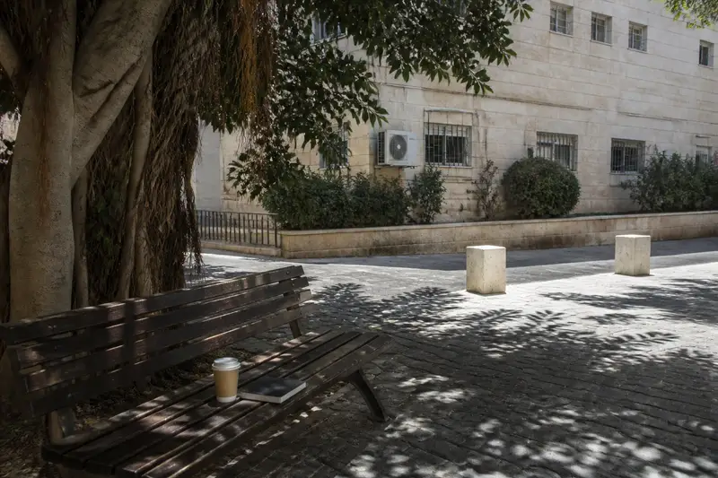 רחבה קטנה מול בניין משרדים בצהריים: ספסל עץ בצל עץ פיקוס, כוס קפה חד פעמית ומחברת סגורה מונחות עליו