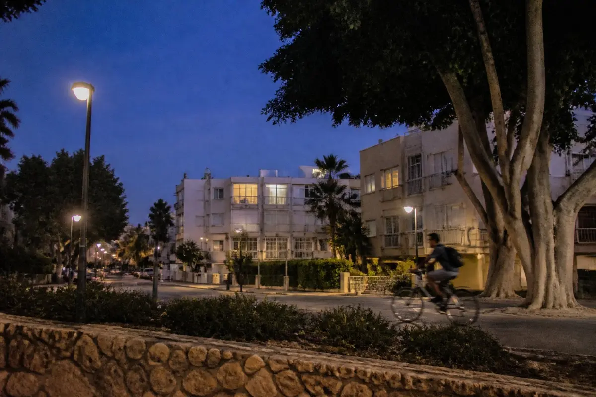 רחוב שקט בשכונת מגורים בשעת דמדומים מוקדמת, פנסי רחוב נדלקים, חלונות מוארים בבנייני דירות נמוכים ורוכב אופניים חולף
