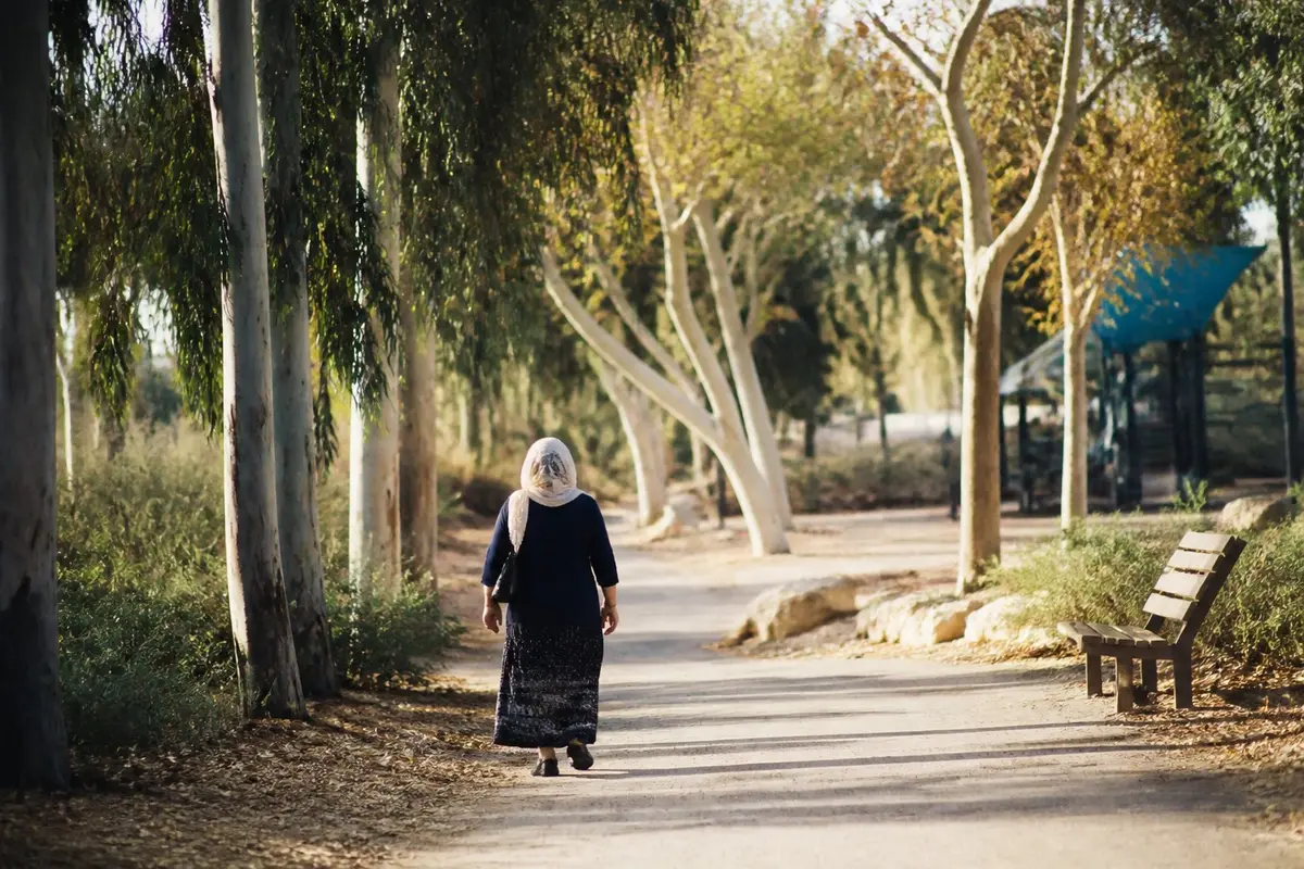 אישה בשנות השישים לחייה עם צעיף הולכת לבד בשביל בפארק עירוני בשעת אחר הצהריים מוקדמת, מצולמת מאחור בין עצים