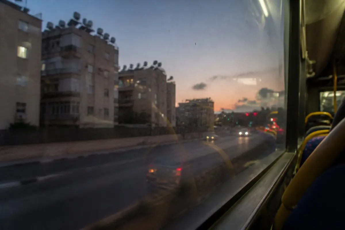נוף מחלון אוטובוס עירוני בשעת דמדומים: רמזורים מטושטשים ובנייני מגורים עם דודי שמש