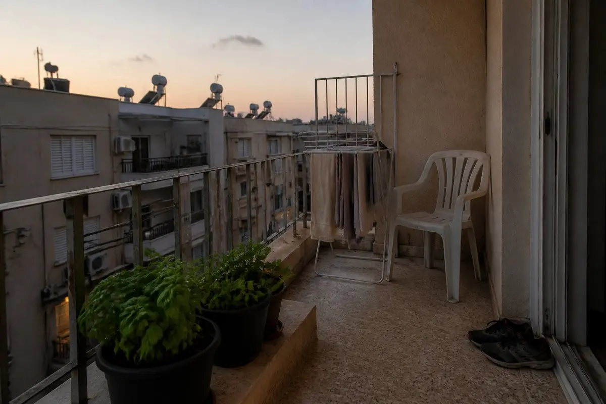 מרפסת קטנה בדירה ישראלית בערב, עם עציצי בזיליקום ונענע, כיסא פלסטיק, מתקן ייבוש כביסה ונעלי ריצה ליד הדלת